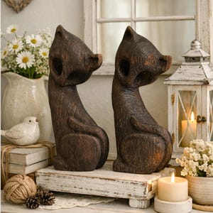 Pode incluir: Duas figuras de gato em madeira castanho-escura com textura de grão de madeira visível, cada uma representada numa posição sentada com as cabeças viradas para o lado. As esculturas apresentam linhas curvas e são exibidas numa base de madeira branca.