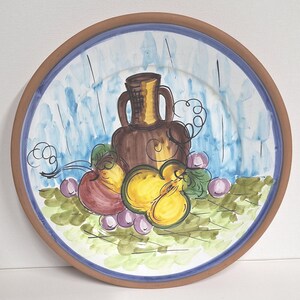 以下が含まれることがあります： 茶色の縁と青と白の内装が施された手描きのセラミックプレート。 プレートには、茶色の花瓶、黄色と赤の果物、紫色のブドウが描かれています。 デザインは黒で縁取られています。