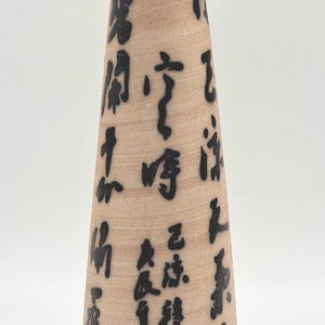 以下が含まれることがあります： 薄茶色のボディと青い縁の陶器の花瓶。花瓶の本体には黒いカリグラフィーが施されています。花瓶の高さは約20cmです。