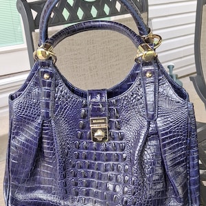 May include: A deep navy blue handbag with a glossy, textured crocodile-embossed finish. The bag features a structured top handle, gold-tone metal hardware, and a central front clasp closure engraved with the brand name BRAHMIN in gold lettering.