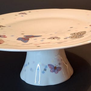 Peut inclure: Présentoir à gâteau en céramique blanche avec un bord festonné et une bordure dorée. L'assiette est décorée de papillons colorés et de petits diamants bleus et gris. La base du présentoir présente également des motifs de papillons.