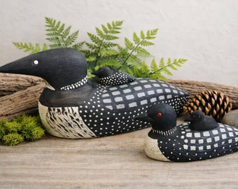 May include: Two carved wooden loon figurines, each featuring a smaller chick resting on its back. The adult loons have black heads, white and black checkered wings, and white spotted bodies. The larger loon has a white eye, while the smaller has a red eye.