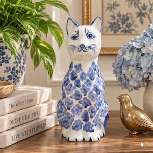 May include: A white ceramic cat figurine featuring a blue floral pattern across its body. The cat has pointed ears, painted facial features including eyes and whiskers, and sits in an upright position. The surface is smooth and glossy.