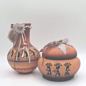 May include: A tan ceramic vase and a matching lidded jar, both decorated with black Kokopelli figures. The vase features a leather tie with red and white beads and feathers. The jar has a rounded lid adorned with feathers and a leather leaf accent.