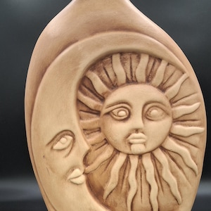 May include: A tan-colored ceramic vase with a narrow neck and a rounded body. The front of the vase features a carved design of the sun and moon. The sun has a face with radiating rays, and the moon has a crescent shape with a face.