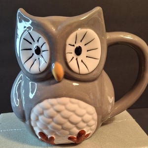 May include: A grey ceramic owl mug with white and black eyes and a brown beak. The owl has a white belly with a scaly pattern and brown feet.