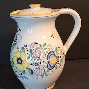 May include: White ceramic pitcher with a lid, featuring a hand-painted floral design in yellow, blue, and green. The pitcher has a curved handle and a yellow rim. A decorative piece for serving beverages.