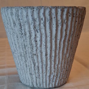 May include: A white concrete planter with a textured, ribbed surface. The planter is a light gray color and has a slightly flared opening.