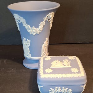 May include: A light blue vase and matching square trinket box. Both pieces feature white bas-relief designs of classical figures, floral garlands, and decorative borders. The vase is approximately 8 inches tall.