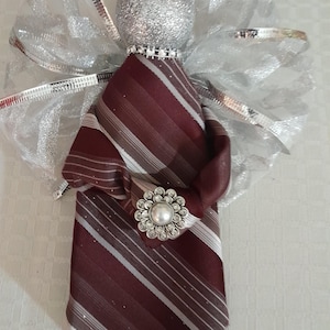 以下が含まれることがあります： バーガンディ、グレー、白のストライプ柄ネクタイで作られた装飾的な天使のオーナメント。天使は、銀色のグリッターボールの頭、銀色のリボンの翼、装飾的なブローチが特徴です。ユニークなホリデーまたは家の装飾アイテム。