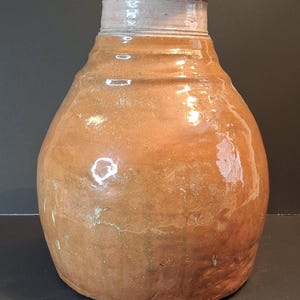 May include: A large, handcrafted ceramic vase with a rounded body and a narrow neck. The vase has a warm, earthy brown colour with a glossy finish. The rim is a lighter shade of brown with a hint of green. The vase is approximately 30 cm tall.
