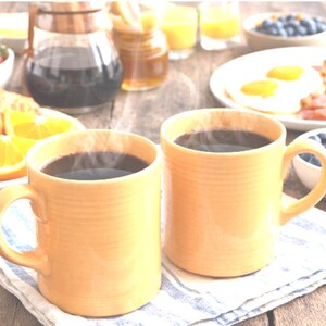 Puede incluir: Dos tazas de cerámica amarilla a juego con detalles acanalados, cada una llena de una bebida oscura humeante. Las tazas están colocadas una al lado de la otra sobre una servilleta de tela a rayas blancas y azules, con un desayuno al fondo.