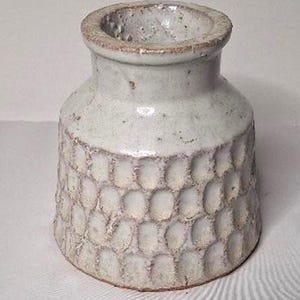 May include: A small, off-white ceramic vase with a textured, honeycomb-like pattern on the lower half. The rim and the pattern's edges have a darker, brownish hue. The vase has a rustic, handmade appearance.