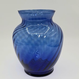 May include: A blue glass vase with a rounded body and a flared rim. The vase has a swirled design, with darker blue accents at the base. The vase is approximately 8 inches tall.