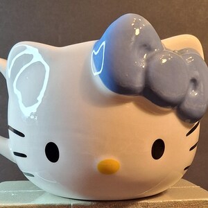 May include: White ceramic mug shaped like a cartoon cat's face, featuring black eyes, whiskers, and a yellow nose. A light blue bow adorns the top. The mug has a handle on the side.