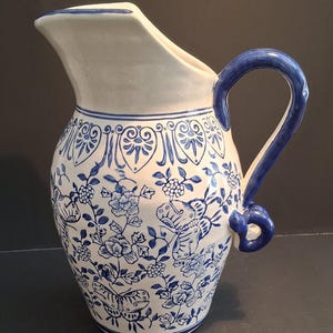 Puede incluir: Una jarra de cerámica blanca con un patrón floral y de mariposas azul. La jarra tiene un pico curvo y un asa grande y arqueada. El diseño incluye flores, hojas y mariposas. El asa y el borde están pintados de azul.