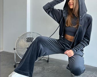 May include: A dark gray velour tracksuit with a zippered hoodie. The model is wearing the tracksuit with white sneakers and is sitting on the floor.