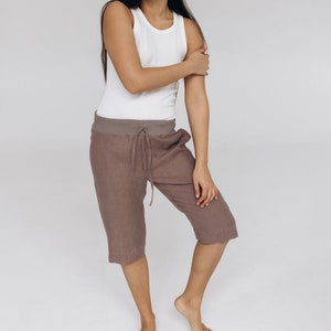 May include: A woman wearing a white vest top and brown linen shorts. She is standing on a white background.