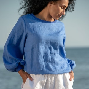Pode incluir: Uma mulher vestindo uma blusa de linho azul claro de mangas compridas e shorts de linho branco. A blusa tem um decote redondo e os shorts têm um caimento relaxado.