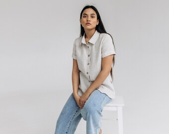 May include: A person wearing a white and brown striped shirt with short sleeves, blue jeans, and white shoes sits on a white stool against a white background.