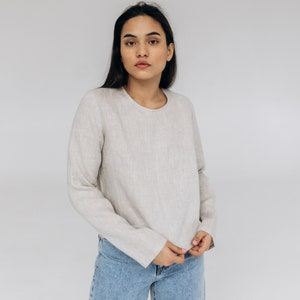 K&ouml;nnte beinhalten: Eine Frau tr&auml;gt ein hellbeiges Langarmshirt und eine blaue Jeans. Das Shirt hat einen runden Ausschnitt und eine lockere Passform.