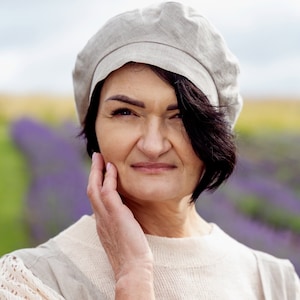 May include: A woman with short dark hair is wearing a light gray linen beret and a light gray linen dress with white lace sleeves. She is standing in a field of lavender.