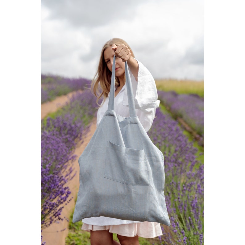 May include: A light blue linen tote bag with a single pocket on the front. The bag is being held by a person in a field of lavender.