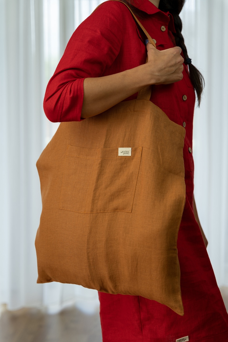 May include: A brown linen tote bag with a large pocket on the front. The bag has a small brown leather tag with the text "The Happy Soul" on it.