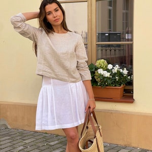 K&ouml;nnte beinhalten: Eine Frau tr&auml;gt ein beiges Leinenoberteil und einen wei&szlig;en Leinenrock. Sie h&auml;lt eine Strohtasche mit braunen Ledergriffen.