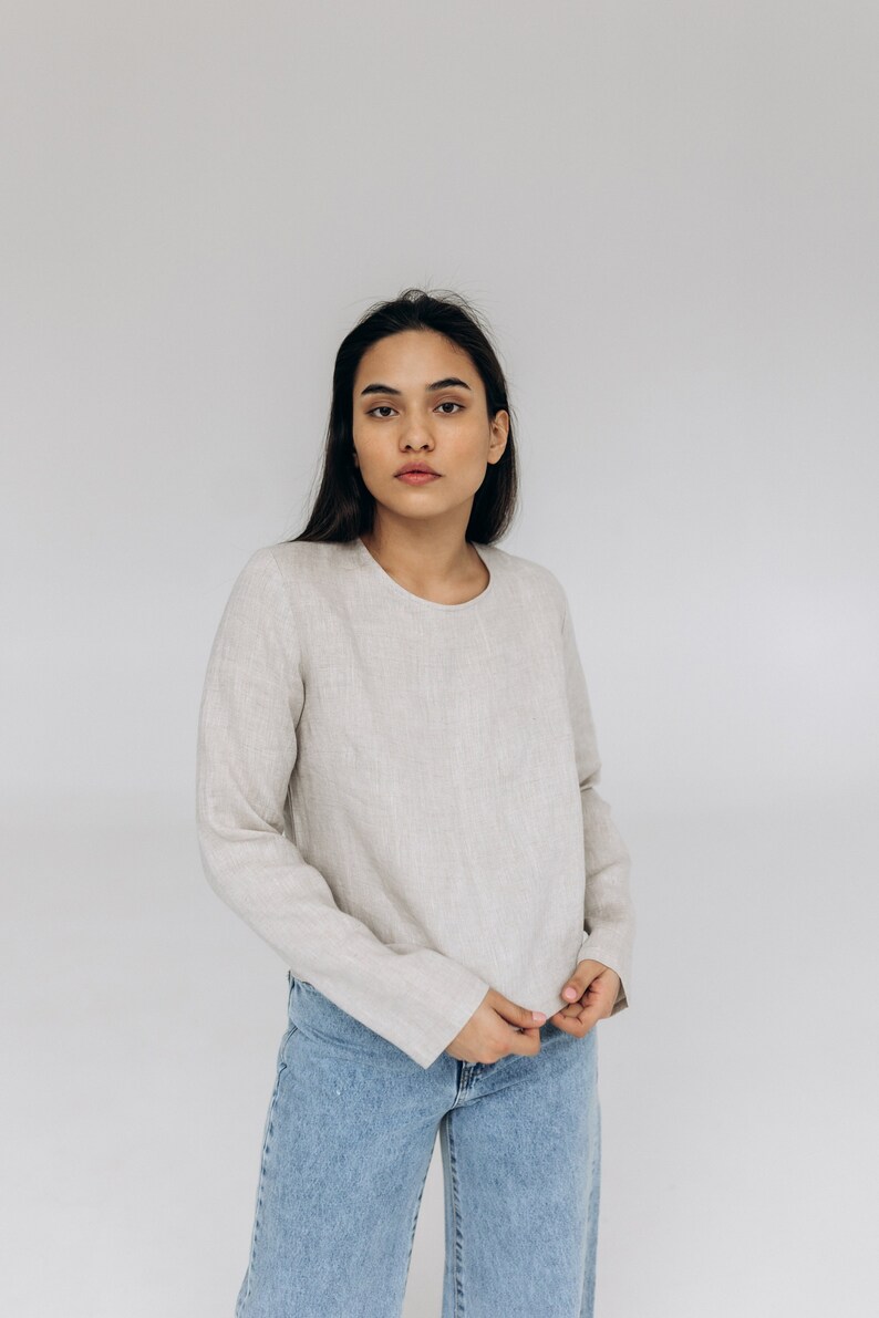 K&ouml;nnte beinhalten: Eine Frau tr&auml;gt ein hellbeiges Langarmshirt und eine blaue Jeans. Das Shirt hat einen runden Ausschnitt und eine lockere Passform.