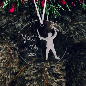 May include: Clear round Christmas ornament with a white silhouette of a person holding a sword. The ornament is personalized with the text "Britt as Fritz 2023".