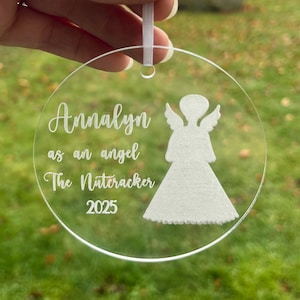 May include: Clear acrylic Christmas ornament with a white angel silhouette and the text "Annalyn as an angel The Nutcracker 2025". The ornament is round and has a white ribbon for hanging.