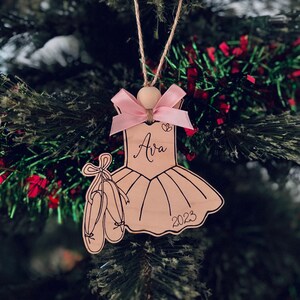May include: Wooden Christmas ornament shaped like a ballerina's dress, with a pink ribbon and a pair of ballet shoes. The name "Ava" and the year "2023" are written on the ornament. The ornament is hanging on a Christmas tree.
