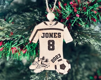 May include: A wooden soccer jersey ornament with the name "JONES" and the number "8" on the front. The ornament also includes a soccer ball and a pair of cleats, and the year "2023" is printed on the bottom.
