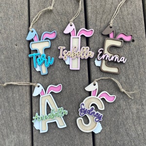 May include: Five wooden ornaments shaped like letters with bunny ears. The letters are T, I, E, A, and S. Each letter has a name written on it: Tori, Isabella, Emma, Annabella, and Sydney. The ornaments are painted in various colors, including pink, blue, brown, and purple.
