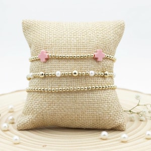 May include: Three gold beaded bracelets are displayed on a beige burlap pillow. One bracelet features pink cross charms, another has pearls, and the third is a simple gold bead design. The bracelets are presented on a wooden surface with scattered pearls.