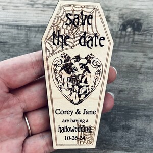 May include: Wooden save the date card shaped like a coffin with a spiderweb design. The card says "Save the date" and features a couple of skeletons in a heart with the text "Corey & Jane are having a halloweeding 10-26-24".