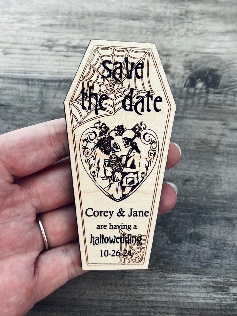 May include: Wooden save the date card shaped like a coffin with a spiderweb design. The card says "Save the date" and features a couple of skeletons in a heart with the text "Corey & Jane are having a halloweeding 10-26-24".