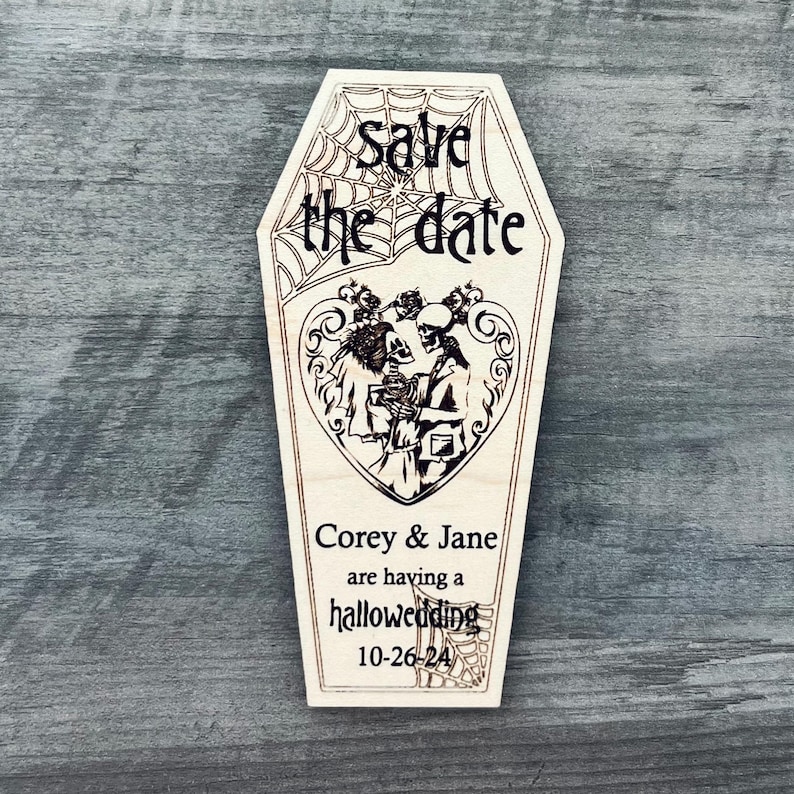 May include: Wooden save the date card shaped like a coffin with a spiderweb design. The card says "Save the date" and features a couple of skeletons dressed as a bride and groom. The text below reads "Corey & Jane are having a halloweeding 10-26-24".