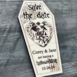 May include: A wooden coffin-shaped save the date card with a spiderweb design and the text "Save the date". The card features a couple of skeletons dressed in wedding attire with the text "Corey & Jane are having a hallowedding 10-26-24".