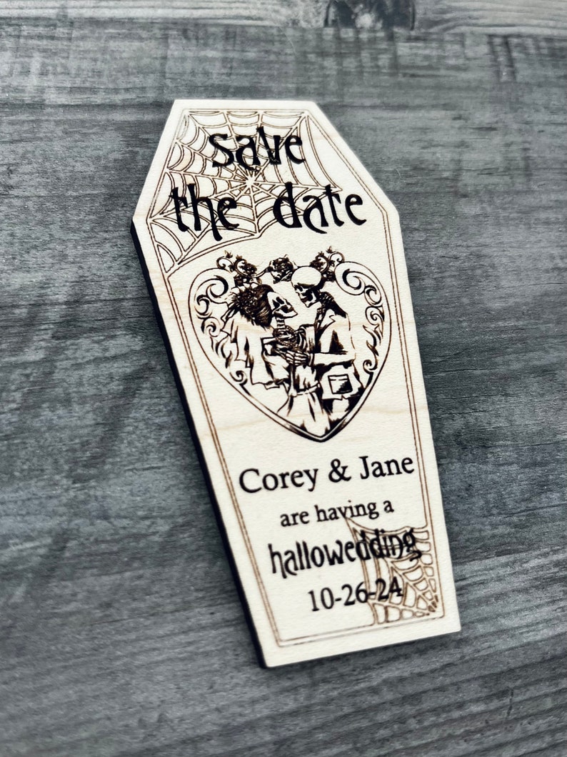 May include: A wooden coffin-shaped save the date card with a spiderweb design and the text "Save the date". The card features a couple of skeletons dressed in wedding attire with the text "Corey & Jane are having a hallowedding 10-26-24".