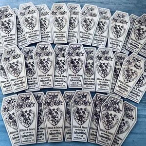 May include: A collection of save the date cards shaped like coffins with a spiderweb design and the text "Save the date" and "Elliot & Mary are having a hallowedding 10-31-25 Middlebury, VT".