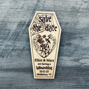 May include: A wooden coffin-shaped save the date card with a skeleton couple in a heart-shaped frame. The text reads "Save the date" with a spiderweb design above it. Below the couple, it says "Elliot & Mary are having a hallowedding 10-31-25 Middlebury, VT".