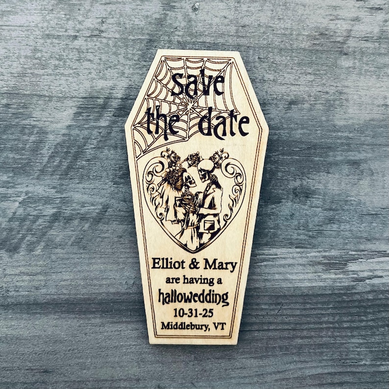 May include: A wooden coffin-shaped save the date card with a skeleton couple in a heart-shaped frame. The text reads "Save the date" with a spiderweb design above it. Below the couple, it says "Elliot & Mary are having a hallowedding 10-31-25 Middlebury, VT".