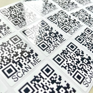 May include: A sheet of white stickers with black and white QR codes. Each sticker has the text "SCAN ME" printed on it.