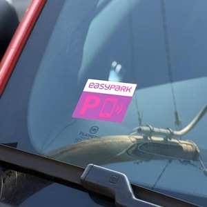Op de afbeelding: Een roze en witte sticker met de tekst "easypark" en een roze "P" met een wit telefoonpictogram erin. De sticker zit op de voorruit van een auto.
