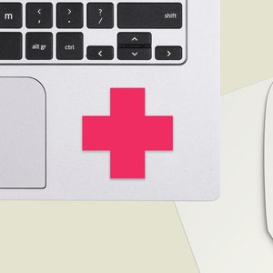 Könnte beinhalten: Ein leuchtend pinkfarbener Pluszeichen-Aufkleber auf einem silbernen Laptop.