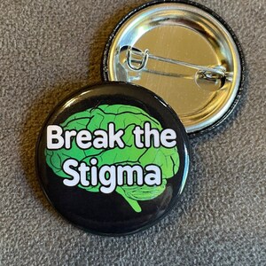 Puede incluir: Un botón negro con un gráfico de cerebro verde y el texto "Break the Stigma" en blanco.