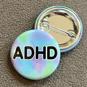Peut inclure: Un bouton bleu et violet tie-dye avec le mot "ADHD" en lettres noires.