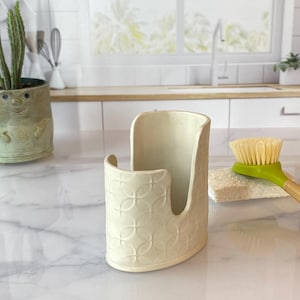 May include: Off-white ceramic sponge holder with a curved design and embossed floral pattern. The holder is next to a green-handled brush and a sponge. The kitchen sink area is in the background.